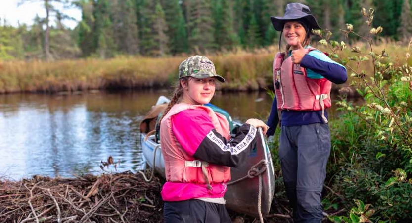 two students smile for a photo while guiding a canoe into water